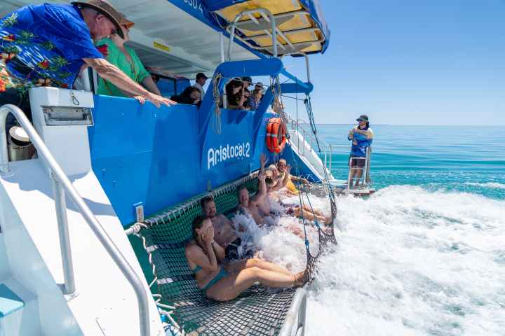 People sit on a net at the back of a boat, enjoying splashes from the sea.