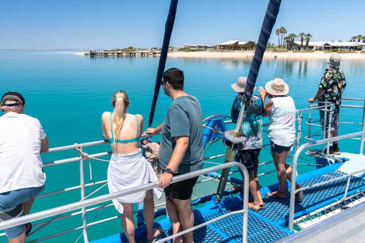 People on a boat watching a coastal beach scene with clear blue water.