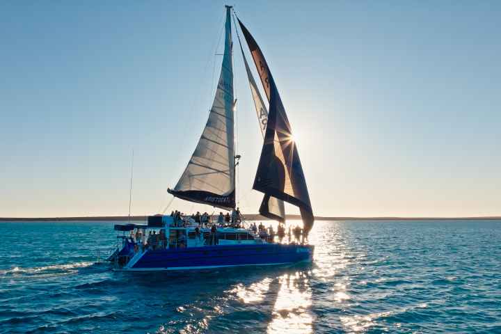 Sailboat with people on deck on calm ocean at sunset, sails partially unfurled.