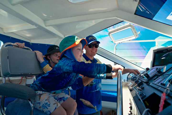 Three people in a boat cabin, one steering while others observe, under a sunny sky.