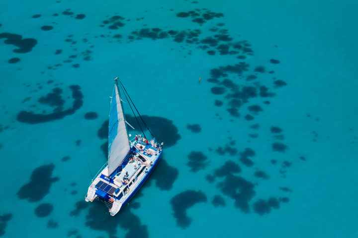 Aerial view of a catamaran sailing on clear turquoise water with patches of coral below.