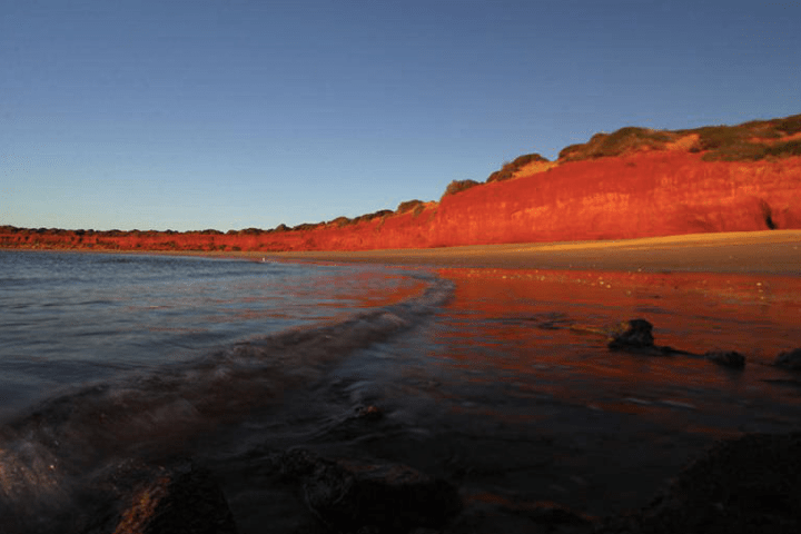 Sunset beach with red cliffs, calm waves, and clear sky.