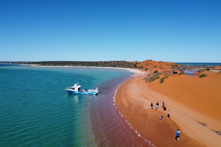 Red sand beach with people walking; nearby a blue boat on turquoise water under clear blue sky.