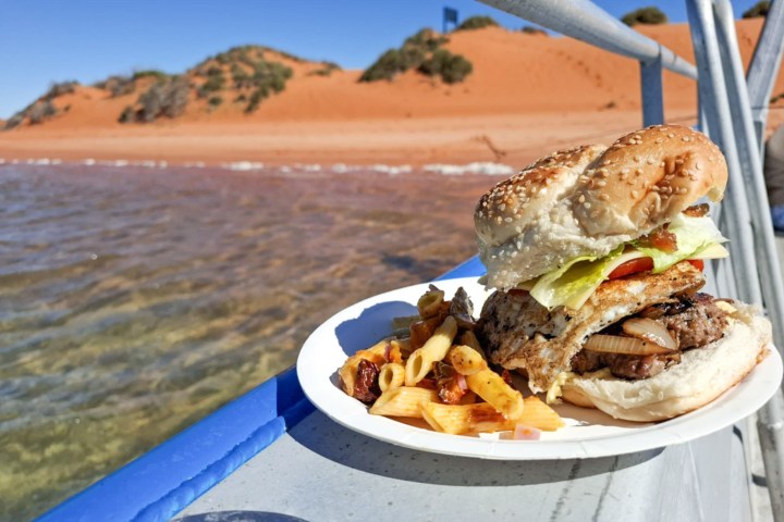 Burger and pasta on plate by water with red sand dunes in background.