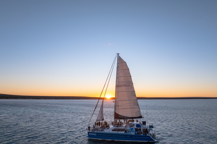 Sailboat on blue water at sunset with people on deck, clear sky.