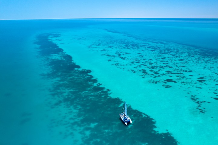 Aerial view of a lone sailboat on a bright turquoise ocean with dark coral patches.