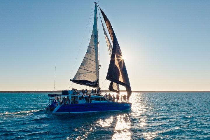 Sailboat with people sailing on calm sea at sunset, sun behind sails.