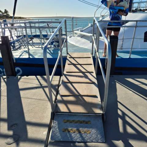 Person on a boat's deck beside a ramp with 'Watch Your Step' sign, overlooking calm sea.