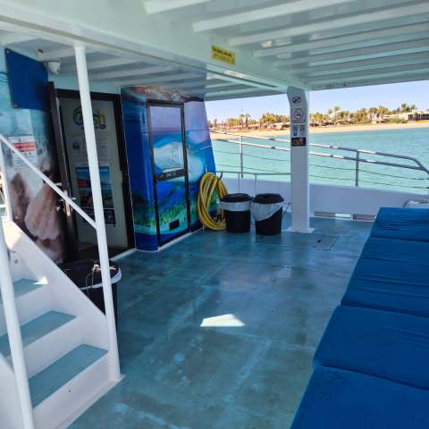 Boat deck with blue seats, stairs, storage area, and view of the sea and coastline in the background.