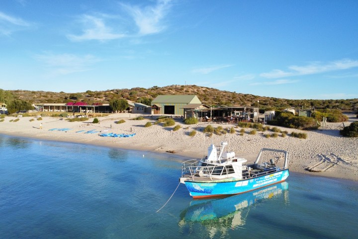 Boat docked by a sandy beach with buildings and trees under blue sky.