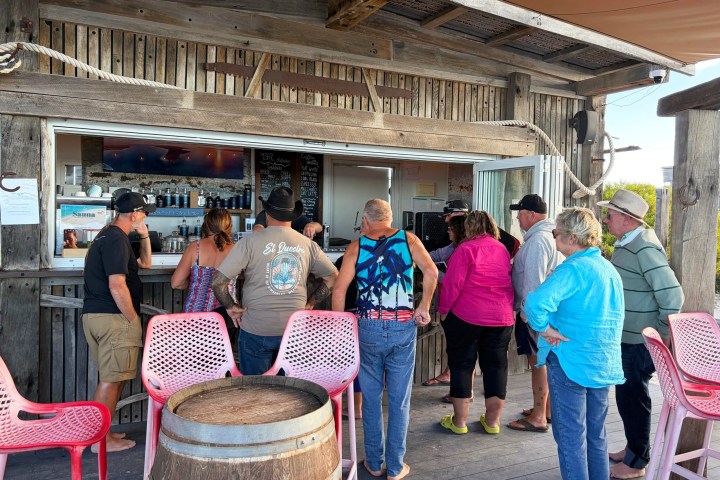 Group of people ordering at a rustic outdoor bar with pink chairs and a barrel table.