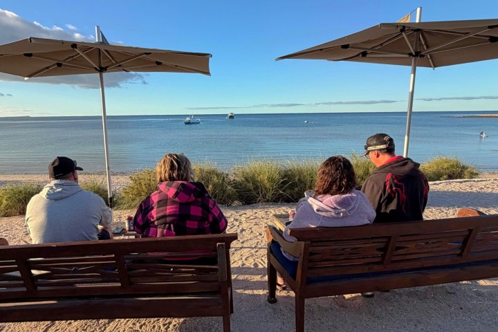 Four people seated on benches by the beach under umbrellas, facing the ocean.
